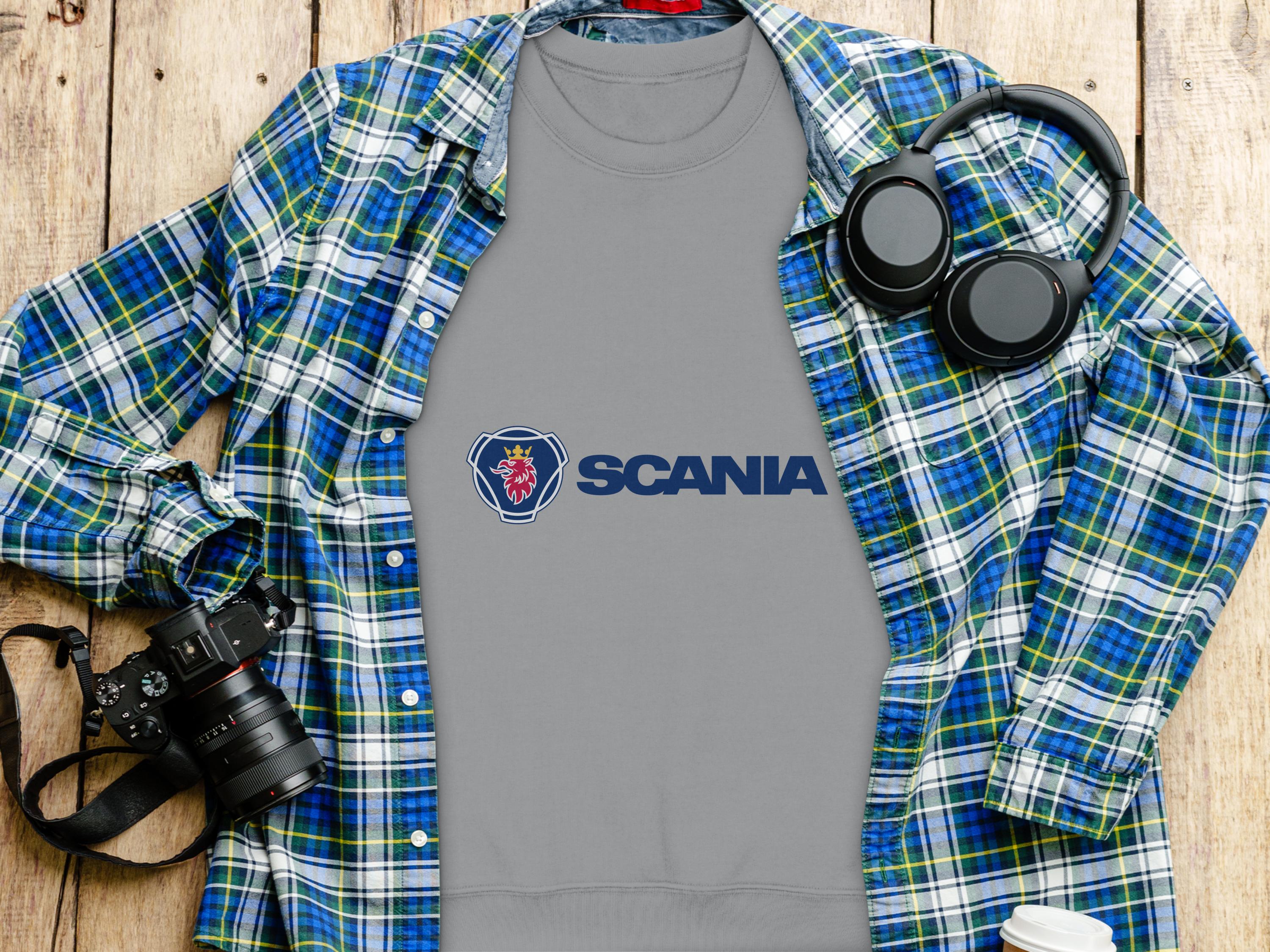 A gray Garment Graphics t-shirt, ideal for casual outings, rests on a wooden surface with a blue and green plaid shirt nearby. Black headphones, a camera with lens, and a white travel mug suggest readiness for adventure—all in unisex sizing.