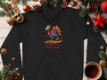 Garment Graphics black hoodie features a cartoon turkey in running shoes with humorous Turkey On The Run text. Pine cones, ornaments, and a cozy coffee cup complement the festive wooden background, making it a quirky hoodie to cherish.