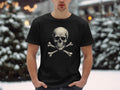 A man stands outdoors in the snow, making a bold fashion statement with a Garment Graphics black graphic T-shirt featuring a skull and crossbones design. Behind him, softly sparkling snow-covered trees add to the wintry charm.