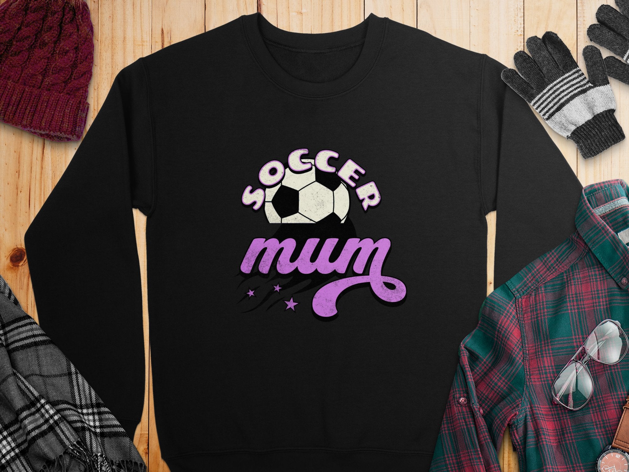 Garment Graphics presents a black sweatshirt with a vintage-style Soccer Mum design in purple and white featuring a soccer ball. Its displayed on wood, accompanied by a maroon knit cap, black and grey gloves, a grey plaid scarf, and a green and red plaid shirt.