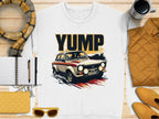 Flat lay of a Garment Graphics white t-shirt with a rally car graphic and YUMP above, surrounded by a pen, notebook, wallet, sunglasses, quilted jacket, scarf, and coffee cup—all in warm yellow and beige tones.