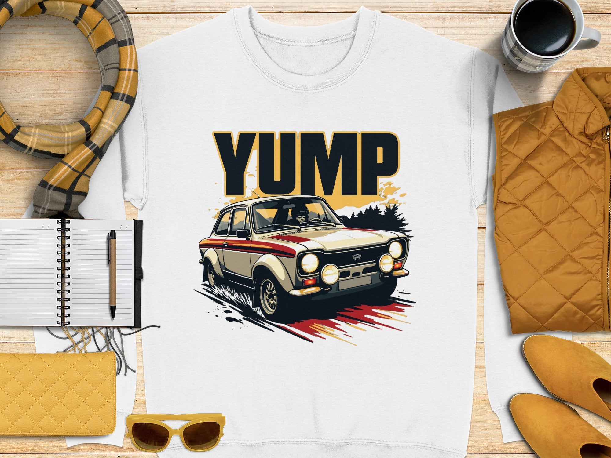 Flat lay of a Garment Graphics white t-shirt with a rally car graphic and YUMP above, surrounded by a pen, notebook, wallet, sunglasses, quilted jacket, scarf, and coffee cup—all in warm yellow and beige tones.