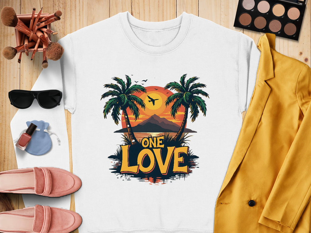 A flat lay features a white sweatshirt from Garment Graphics adorned with the One Love Sunset design, showcasing palm trees and a serene beach. Complementing it are sunglasses, a mustard yellow jacket, makeup brushes, an eyeshadow palette, pink shoes, nail polish, and a chic pouch.