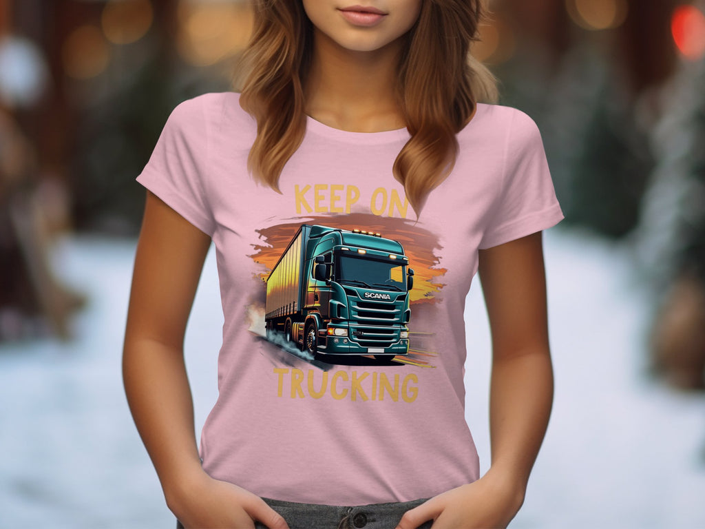 A woman wears a Garment Graphics light pink T-shirt featuring a truck graphic and the slogan KEEP ON TRUCKING, ideal for trucking fans. Blurred trees and snow in the background emphasize the winter setting.
