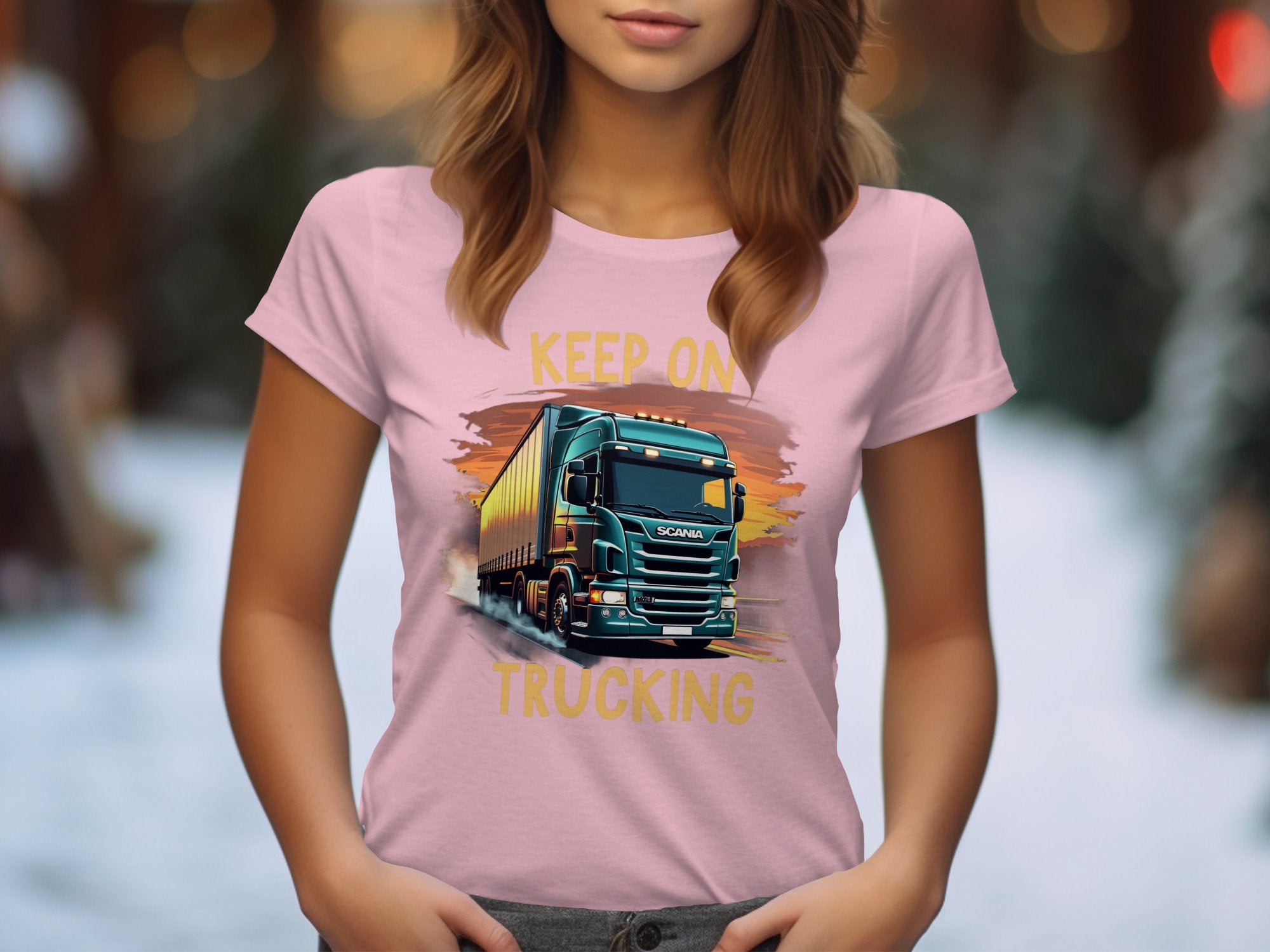 A woman wears a Garment Graphics light pink T-shirt featuring a truck graphic and the slogan KEEP ON TRUCKING, ideal for trucking fans. Blurred trees and snow in the background emphasize the winter setting.