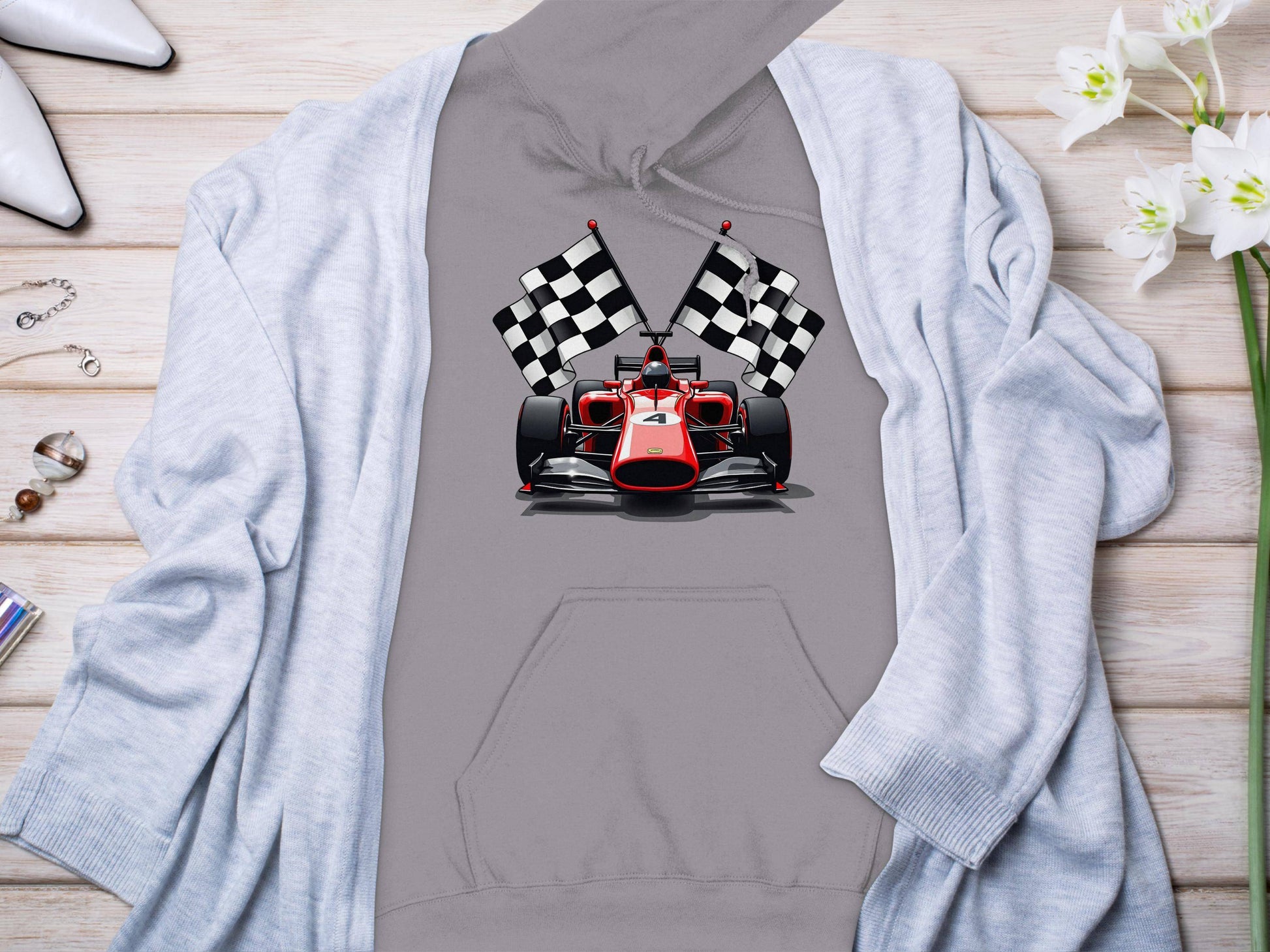 A Garment Graphics gray hoodie featuring a racing car and checkered flags is displayed on wood, surrounded by white high heels, sparkling jewelry, delicate white flowers, and a light blue cardigan elegantly draped over it.