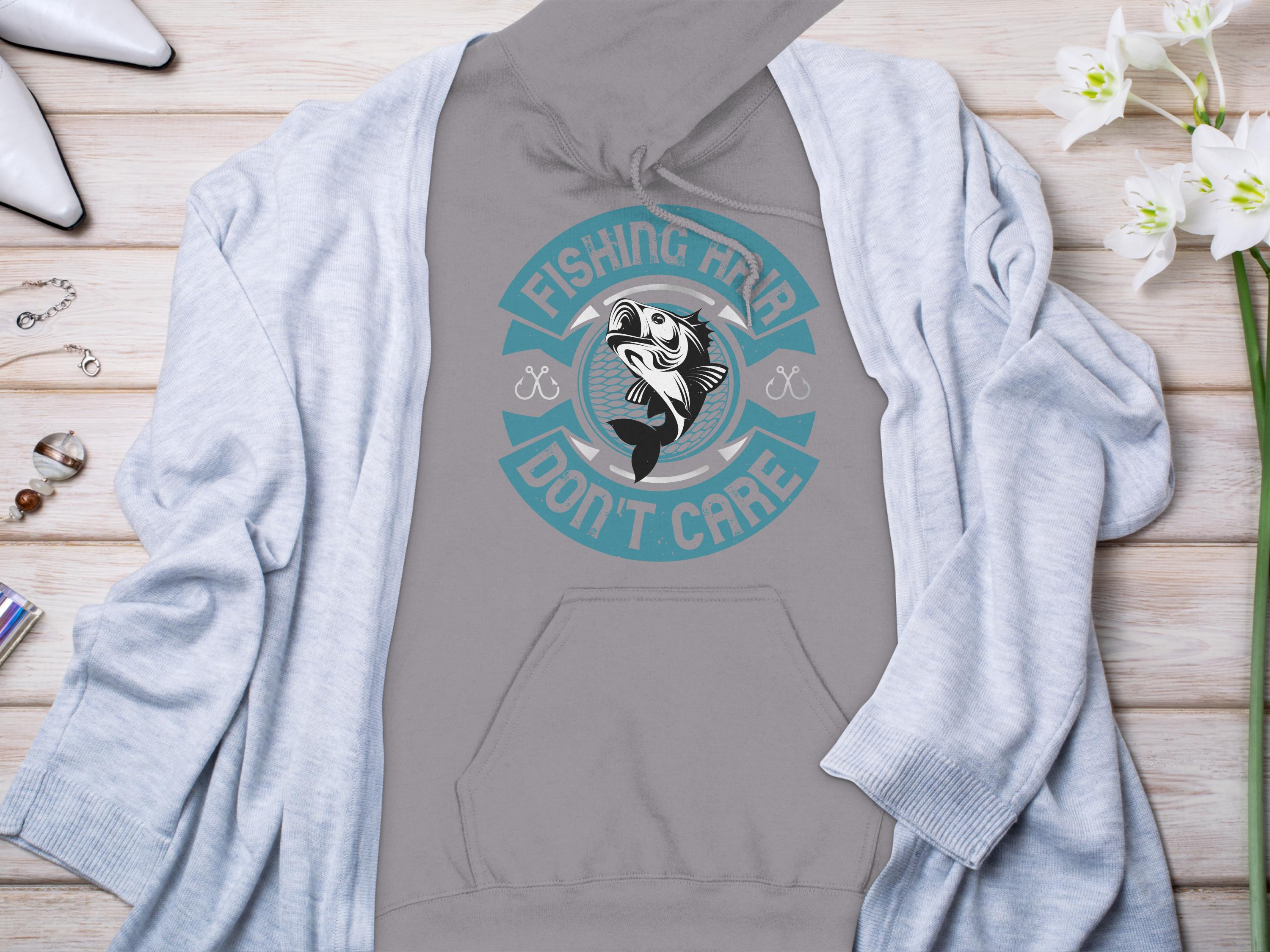 A gray Fishing Hair Dont Care hoodie by Garment Graphics, featuring a fish graphic, sits on light wood. Paired with a light blue cardigan, white flowers, white shoes, bracelet, and earrings, it creates the perfect look for any stylish angler.