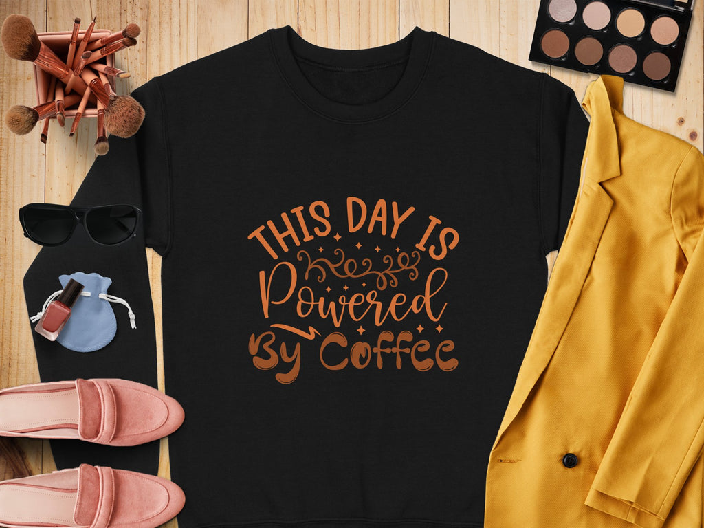 Flat lay of Garment Graphics graphic sweatshirt with This day is powered by coffee in brown, surrounded by makeup brushes, eyeshadow palette, sunglasses, lipstick, red nail polish, pink shoes, blue pouch, and mustard yellow blazer on wood—a playful design for coffee lovers.