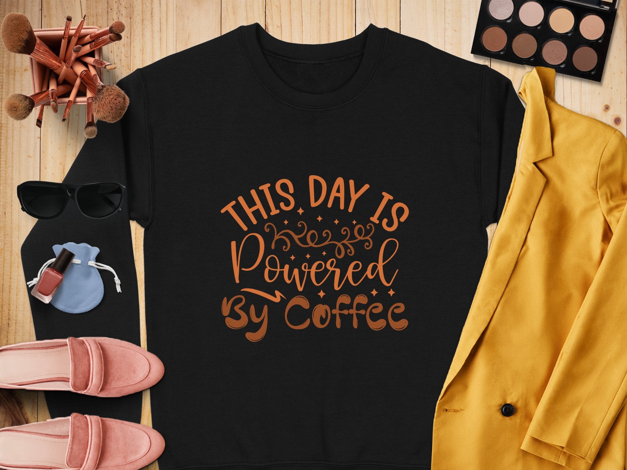 Flat lay of Garment Graphics graphic sweatshirt with This day is powered by coffee in brown, surrounded by makeup brushes, eyeshadow palette, sunglasses, lipstick, red nail polish, pink shoes, blue pouch, and mustard yellow blazer on wood—a playful design for coffee lovers.