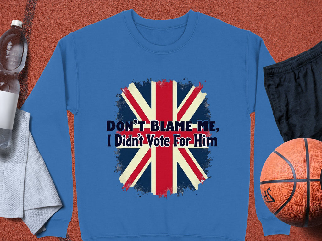 A blue Garment Graphics sweatshirt with a bold Union Jack and DONT BLAME ME, I Didnt Vote For Him design rests next to a water bottle, towel, basketball, and shorts.