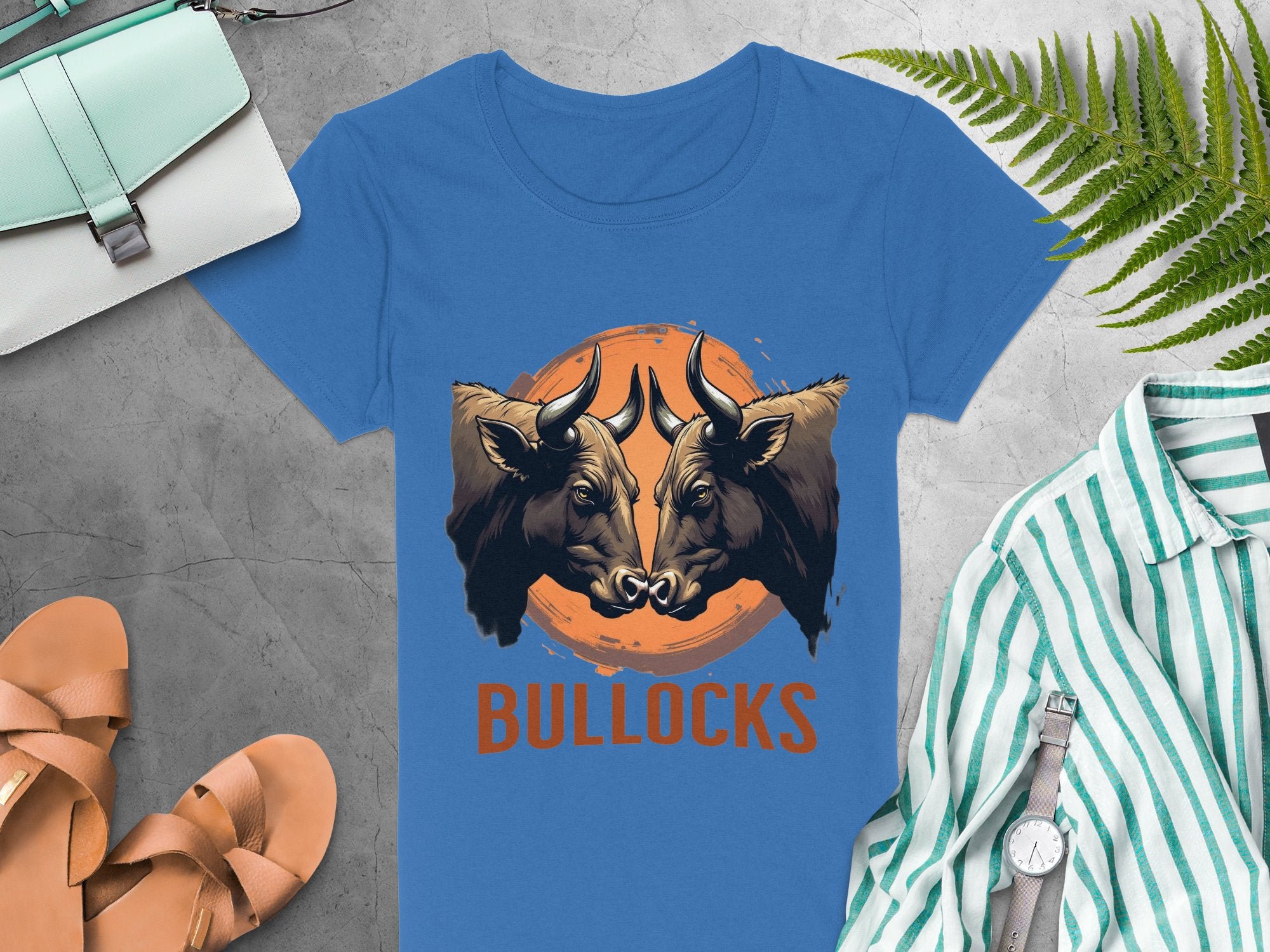 A blue Garment Graphics t-shirt with bold bull illustrations and BULLOCKS lettering, perfect for casual wear, is displayed on concrete alongside a green striped shirt, sandals, a watch, a light blue bag, and green leaves.