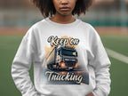 Someone wears a unisex Garment Graphics sweatshirt with a large truck design and Keep on Trucking text. The blurry outdoor background suggests an adventurous vibe, enhancing the casual style.