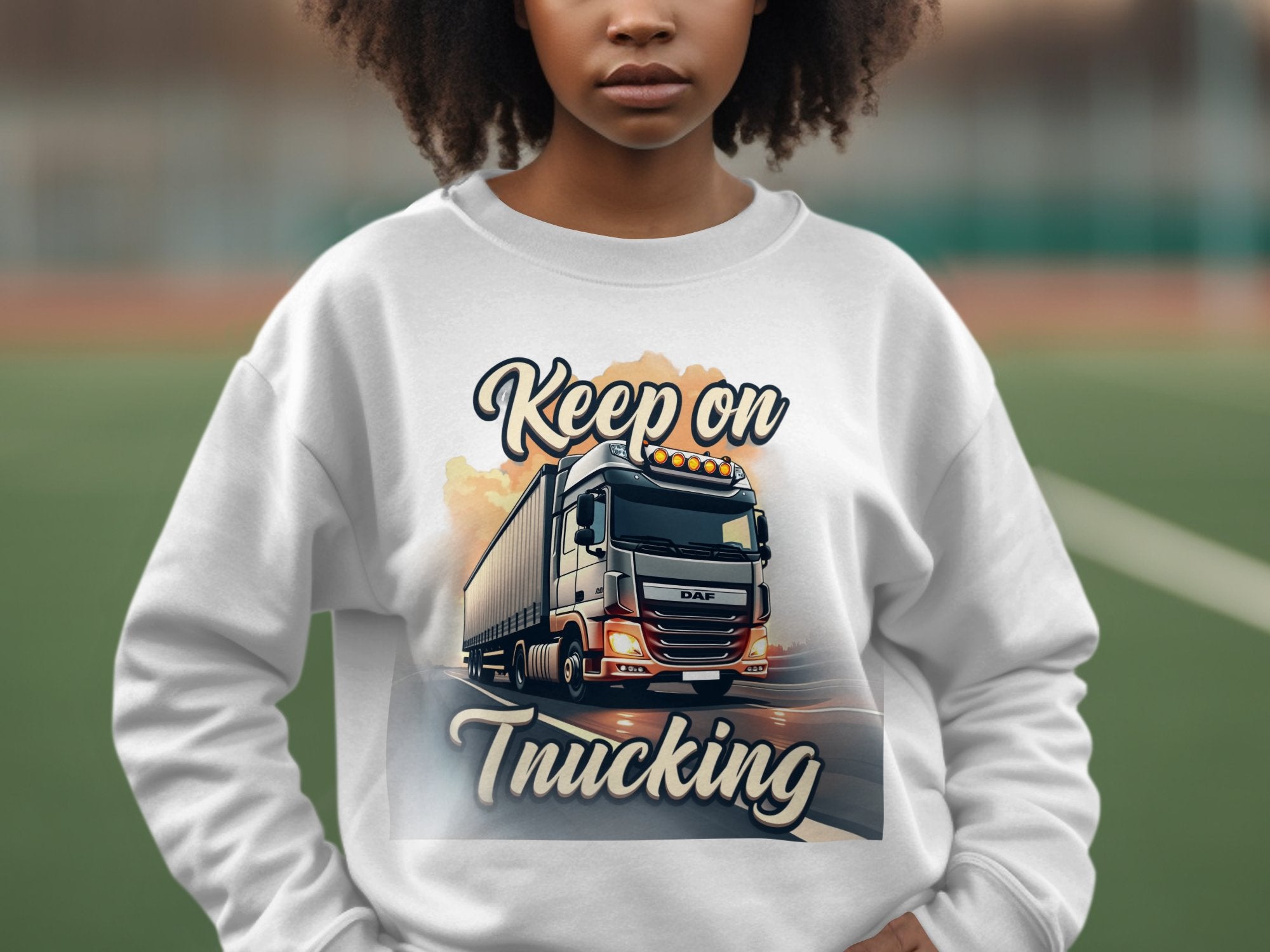 Someone wears a unisex Garment Graphics sweatshirt with a large truck design and Keep on Trucking text. The blurry outdoor background suggests an adventurous vibe, enhancing the casual style.