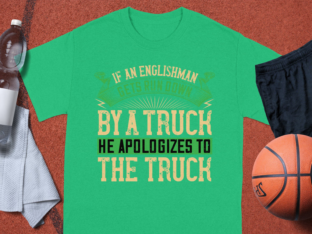 A green T-shirt by Garment Graphics humorously states, “If an Englishman gets run down by a truck, he apologizes to the truck.” It rests on a brown surface beside a basketball, black shorts, a water bottle, and a white towel.