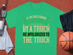 A green T-shirt by Garment Graphics humorously states, “If an Englishman gets run down by a truck, he apologizes to the truck.” It rests on a brown surface beside a basketball, black shorts, a water bottle, and a white towel.