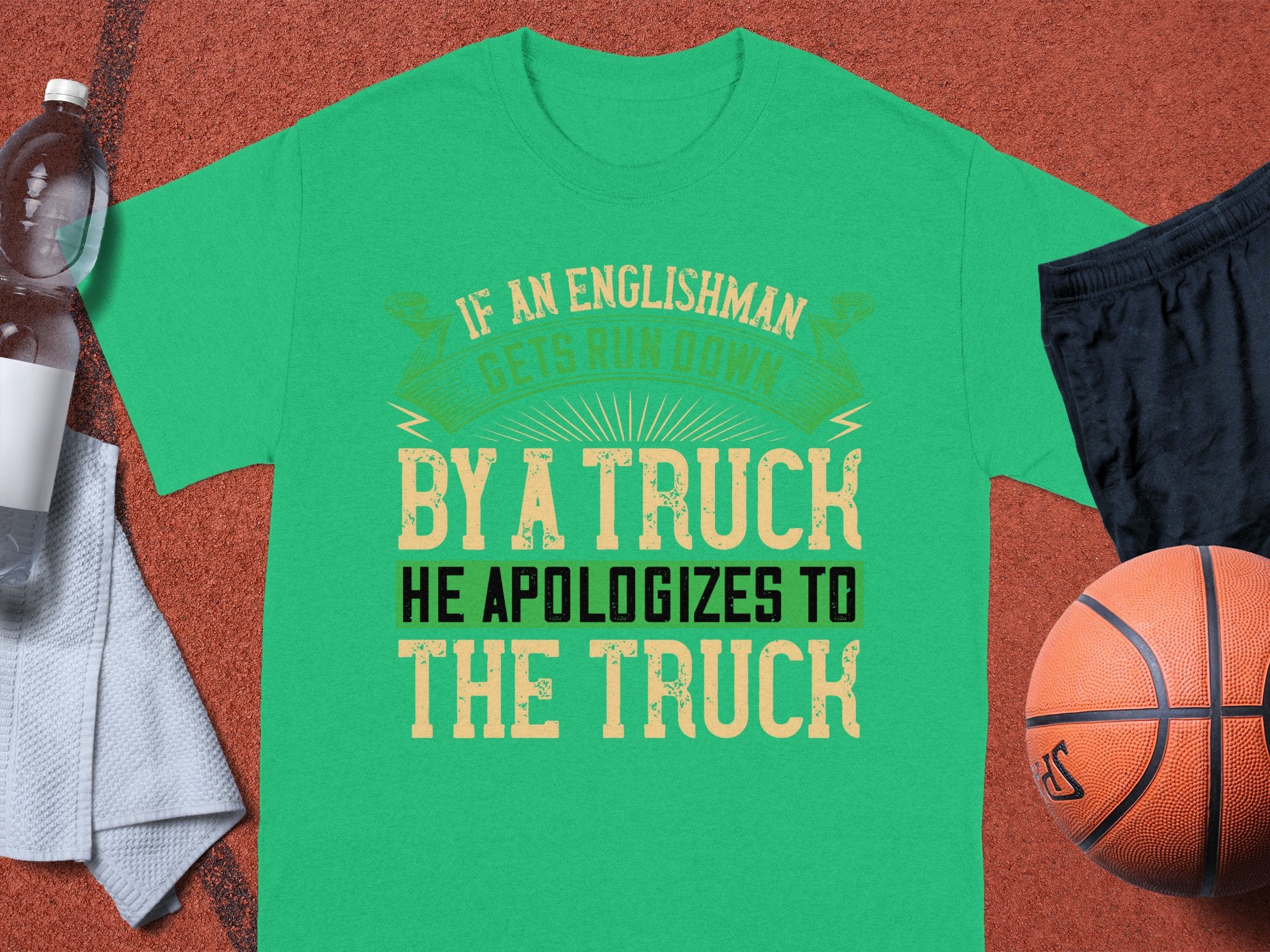 A green T-shirt by Garment Graphics humorously states, “If an Englishman gets run down by a truck, he apologizes to the truck.” It rests on a brown surface beside a basketball, black shorts, a water bottle, and a white towel.