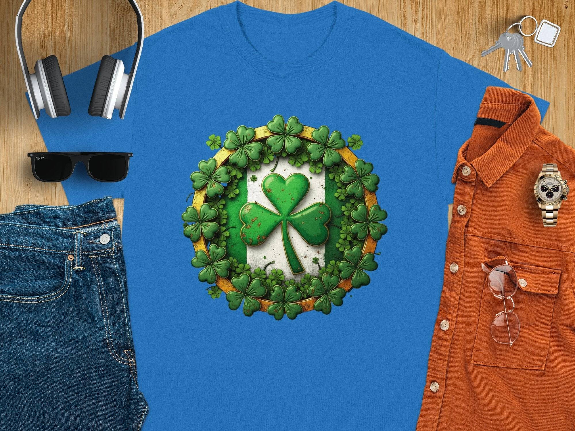 The Garment Graphics blue tee features a vibrant green shamrock clover design for St. Patricks Day, styled on wood with headphones, sunglasses, jeans, an orange shirt, keys, and a gold watch.