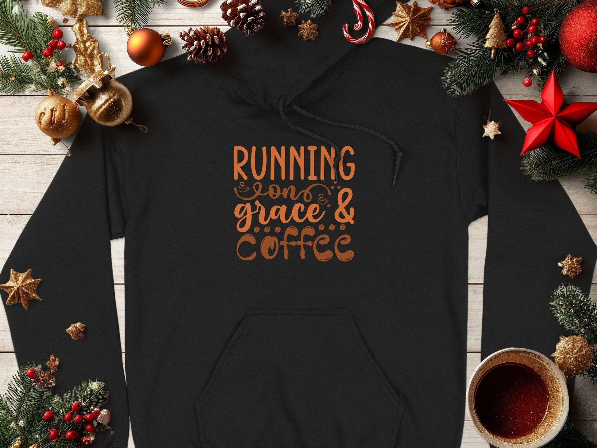 A classic fit black hoodie from Garment Graphics with Running on Grace & Coffee in orange lies on a wooden surface. Surrounded by Christmas decorations like pine cones, baubles, stars, and a steaming cup of coffee, this medium-heavy fabric piece exudes festive coziness.