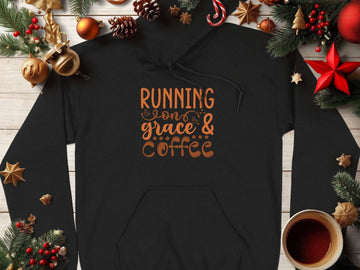 A classic fit black hoodie from Garment Graphics with Running on Grace & Coffee in orange lies on a wooden surface. Surrounded by Christmas decorations like pine cones, baubles, stars, and a steaming cup of coffee, this medium-heavy fabric piece exudes festive coziness.