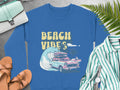 A blue sweatshirt from Garment Graphics showcases a classic car and surfboard with BEACH VIBES, surrounded by palm trees and waves. Styled with sandals, a striped shirt, a watch, purse, and ferns on gray, its ideal for retro vibes and beach fun.