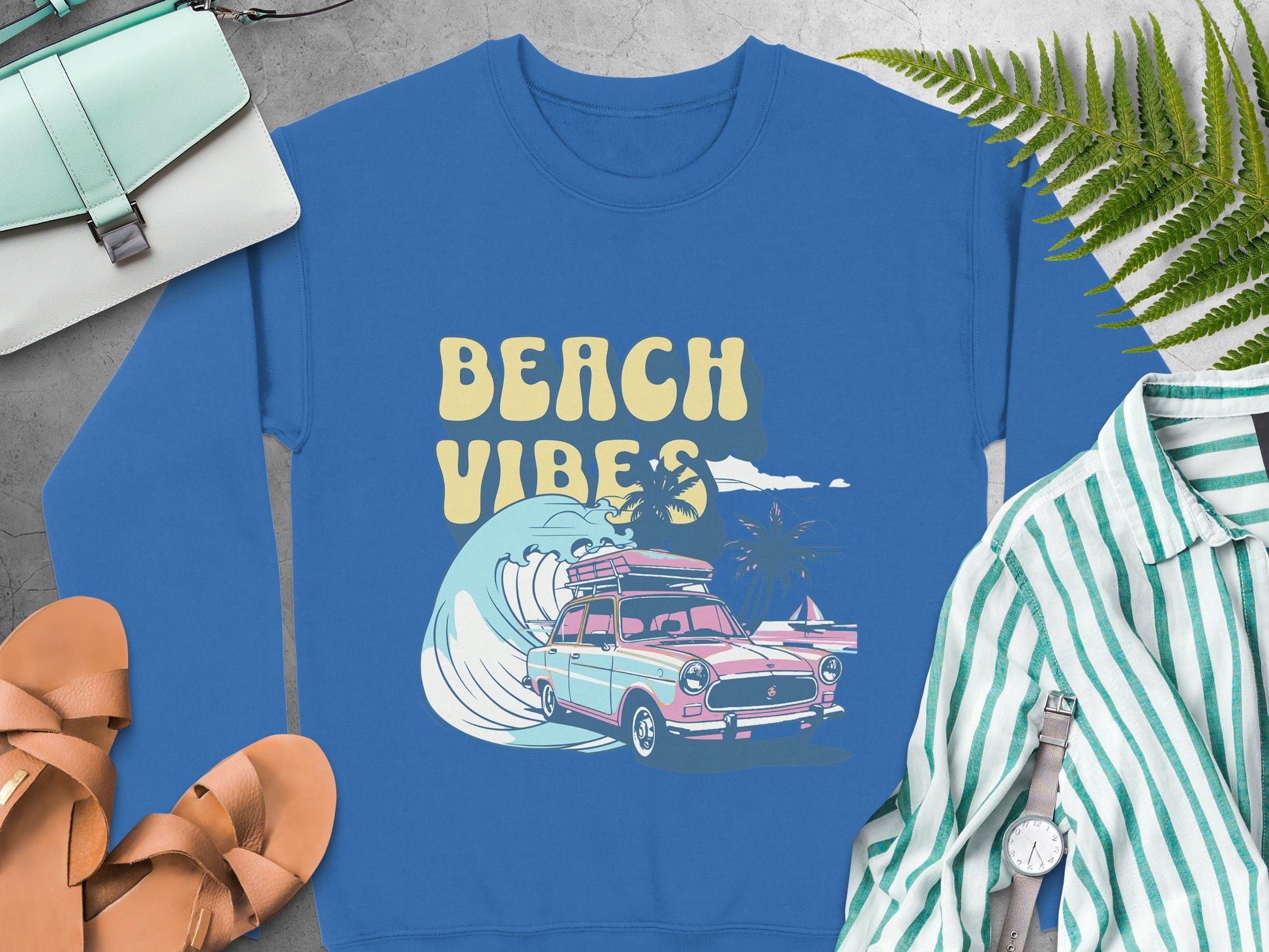 A blue sweatshirt from Garment Graphics showcases a classic car and surfboard with BEACH VIBES, surrounded by palm trees and waves. Styled with sandals, a striped shirt, a watch, purse, and ferns on gray, its ideal for retro vibes and beach fun.