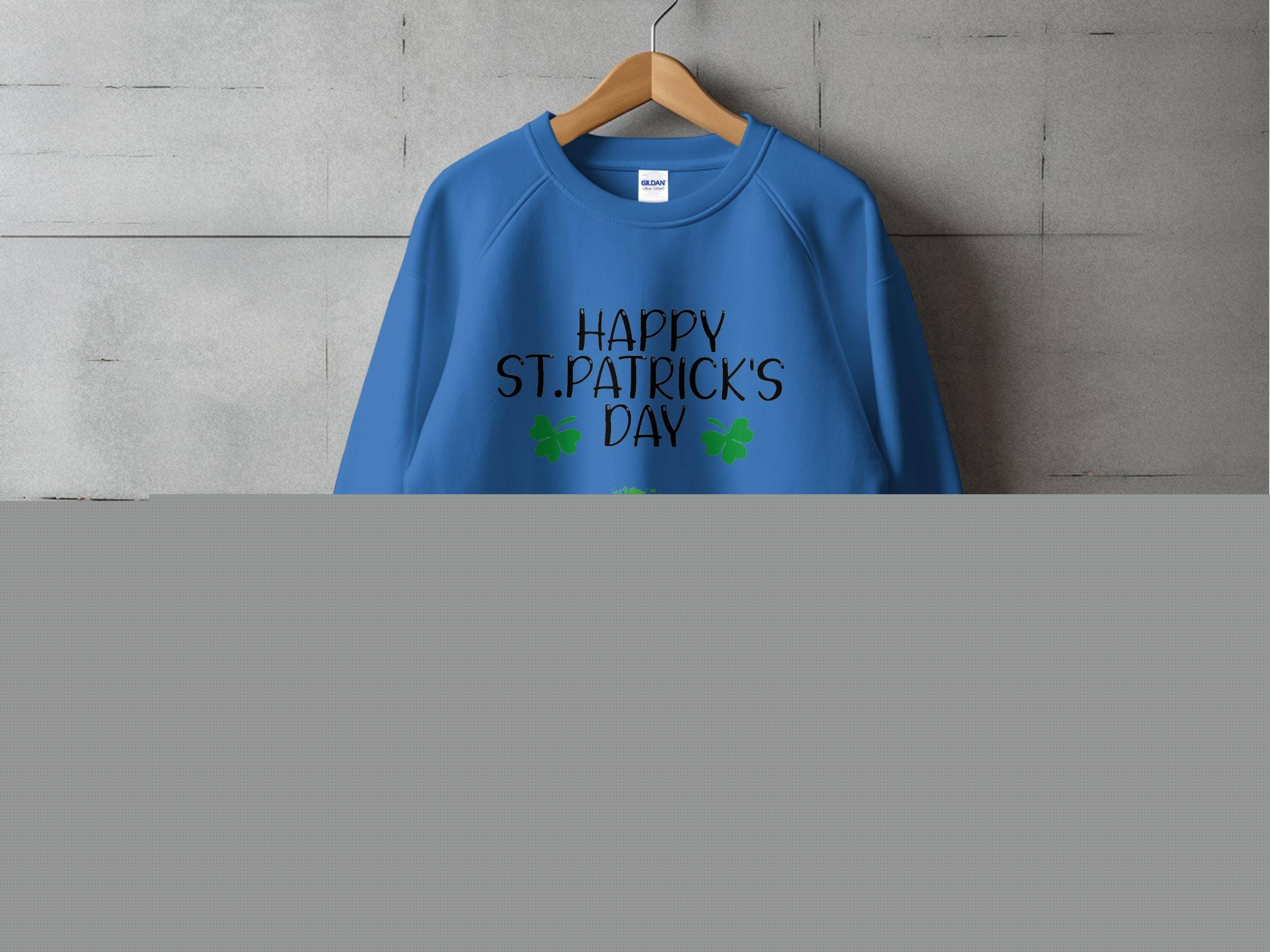 Displayed on a wooden hanger against a gray wall, the blue sweatshirt by Garment Graphics features Happy St. Patricks Day in bold black text, complemented by two green shamrocks and a playful clover kiss below, perfectly capturing the festive spirit.