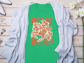 A green Garment Graphics t-shirt featuring a cowboy on horseback and the text Howdy Rodeo, ideal for rodeo fans, is displayed. It’s partially covered by a gray cardigan on a wooden surface, surrounded by white flowers and various accessories.