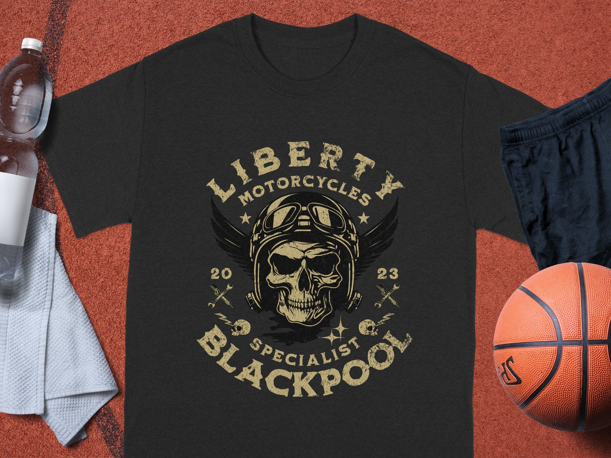 The black t-shirt by Garment Graphics displays a vintage design of a skull with helmet and goggles, bordered by Liberty Motorcycles Specialist Blackpool 2023. Ideal for motorcycle fans, it’s showcased alongside a water bottle, towel, basketball, and black shorts on a textured backdrop.