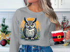 In a festive kitchen, someone holds a Santa-themed mug and wears a Garment Graphics gray sweatshirt featuring an owl on an orange background. Its unisex sizing ensures comfort for all. Decorations like a small tree and ornaments enhance the holiday atmosphere.