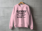 A light pink unisex sweatshirt by Garment Graphics hangs on a wooden hanger against a gray concrete wall. It humorously displays Cleverly disguised as a responsible adult in black text with decorative arrows, adding playfulness to any wardrobe.