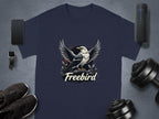 A navy blue, 100% cotton t-shirt from Garment Graphics flaunts a bold eagle graphic with spread wings and the word Freebird below. Its accompanied by a smartphone, smartwatch, fitness tracker, dumbbells, and black sneakers.