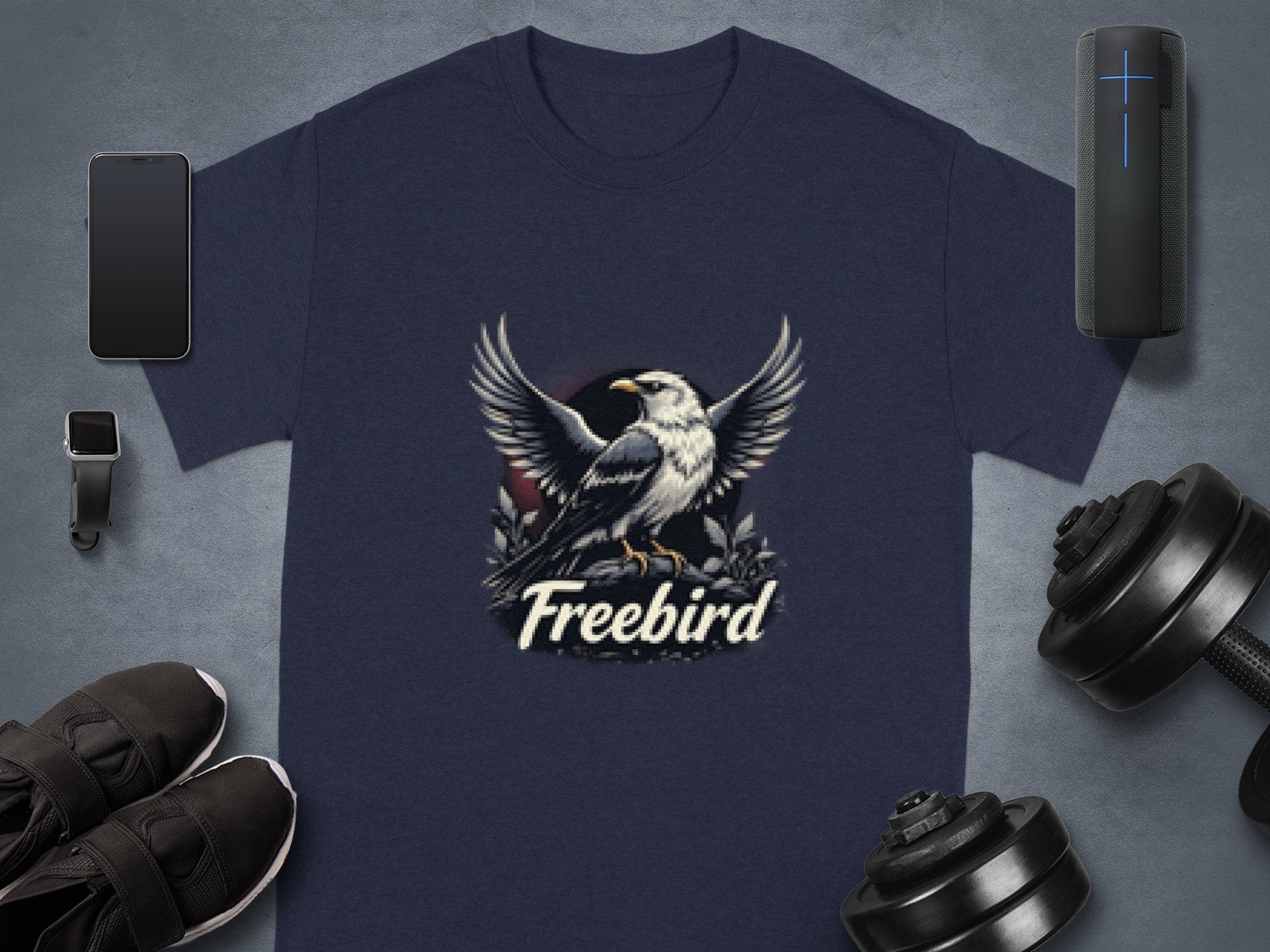 A navy blue, 100% cotton t-shirt from Garment Graphics flaunts a bold eagle graphic with spread wings and the word Freebird below. Its accompanied by a smartphone, smartwatch, fitness tracker, dumbbells, and black sneakers.