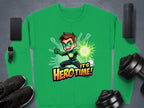 A green sweatshirt by Garment Graphics features a spiky brown-haired cartoon character in a superhero pose with a green mask and gloves, Its Hero Time! text. This unisex graphic print sweatshirt is styled alongside gadgets, shoes, and dumbbells on a gray surface.