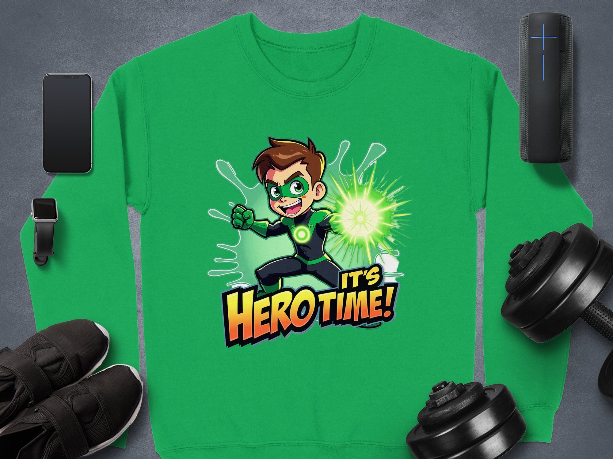 A green sweatshirt by Garment Graphics features a spiky brown-haired cartoon character in a superhero pose with a green mask and gloves, Its Hero Time! text. This unisex graphic print sweatshirt is styled alongside gadgets, shoes, and dumbbells on a gray surface.