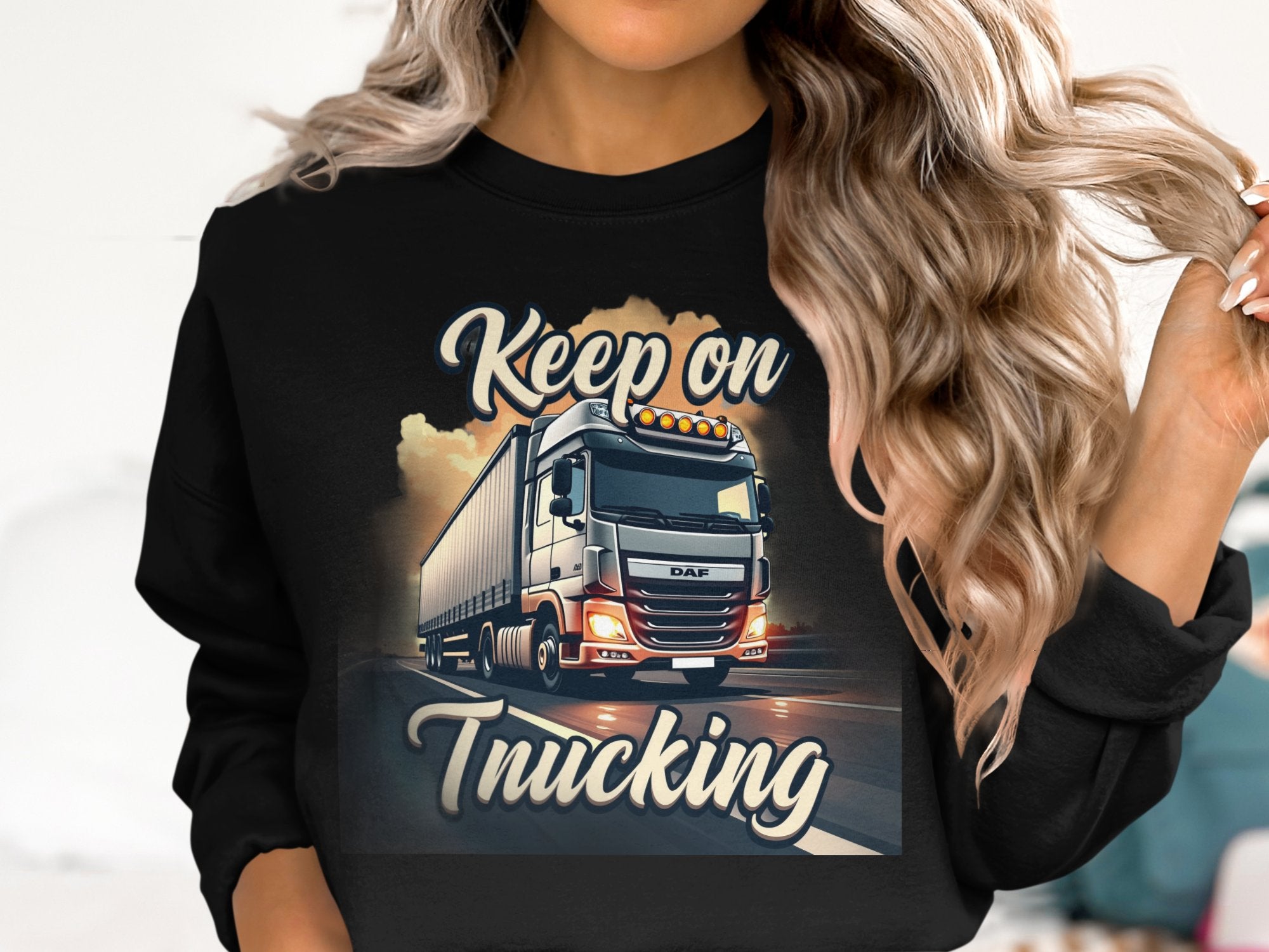 A woman with long, wavy blonde hair wears a black Garment Graphics sweatshirt featuring a truck and Keep On Trucking text. This stylish piece offers unisex sizing for maximum comfort.
