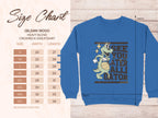 This blue Garment Graphics sweatshirt showcases a cartoon alligator holding a basketball with See you later alligator text. It features unisex Gildan 18000 sizing from small to 5XL.