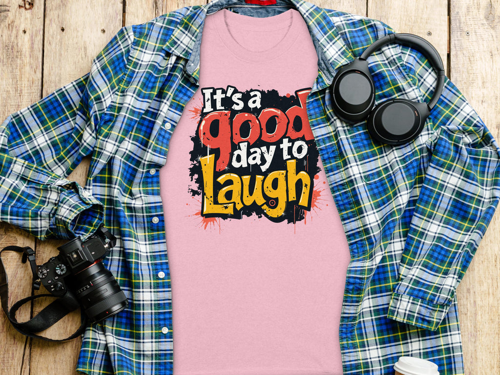 Garment Graphics presents a pink Its a Good Day to Laugh t-shirt with its joyful slogan resting on wood. A blue plaid shirt is artfully spread around, surrounded by accessories: a camera and headphones on the left and a cup to the right.