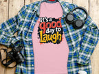 Garment Graphics presents a pink Its a Good Day to Laugh t-shirt with its joyful slogan resting on wood. A blue plaid shirt is artfully spread around, surrounded by accessories: a camera and headphones on the left and a cup to the right.