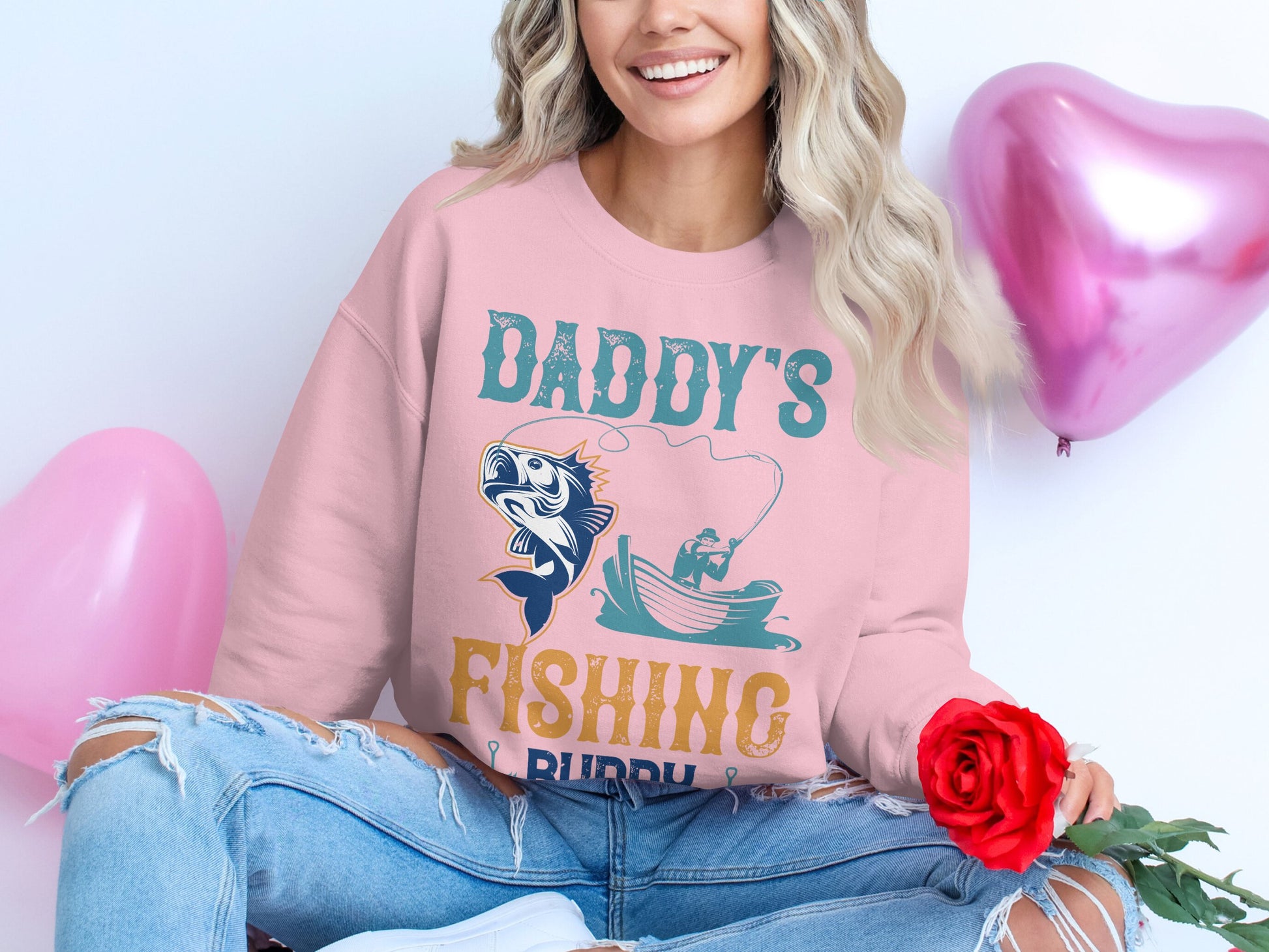 A woman with long, wavy hair smiles while sitting cross-legged. She wears a Garment Graphics pink Daddys Fishing Buddy sweatshirt and ripped jeans, holding a red rose. Pink heart-shaped balloons float in the background, capturing joyful moments for fishing enthusiasts.