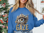 A person stands indoors by a decorated Christmas tree, gently holding their hair and wearing a blue Garment Graphics unisex graphic sweatshirt featuring an ornate eye design with GAZE printed below, offering a comfortable fit for everyone.