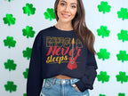 A woman with long hair stands against a white wall with green shamrock cutouts, wearing Garment Graphics trendy sweatshirt featuring Music Never Sleeps Guitar text and a red guitar graphic. She pairs it perfectly with denim shorts—a must-have for music lovers.