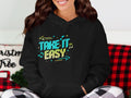 Wearing a Garment Graphics black hoodie with Take It Easy in bright colors, musical notes, and arrows, they sit comfortably with a red and black plaid blanket and a pillow featuring the word Christmas.