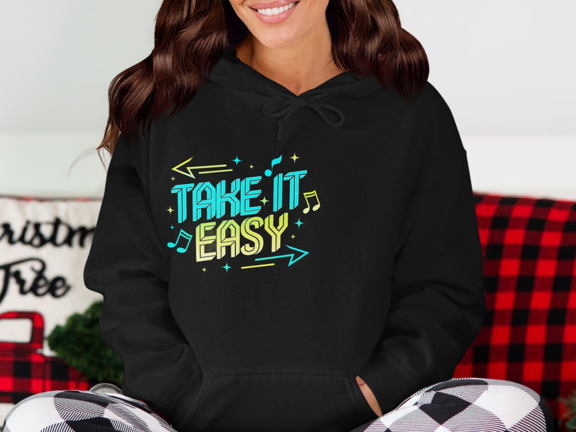 Wearing a Garment Graphics black hoodie with Take It Easy in bright colors, musical notes, and arrows, they sit comfortably with a red and black plaid blanket and a pillow featuring the word Christmas.