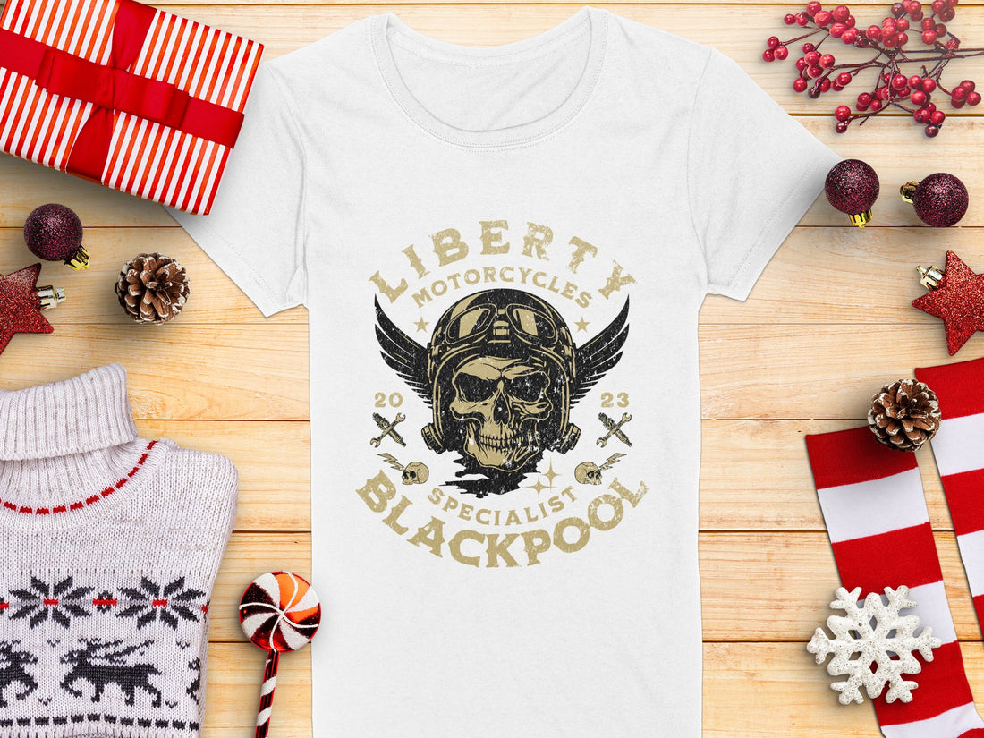 A Blackpool-themed white t-shirt from Garment Graphics features a skull with wings, pilot helmet, goggles, and Liberty Motorcycles Specialist Blackpool 2023. Its artistically displayed on a wooden surface with festive decor and a cozy sweater.
