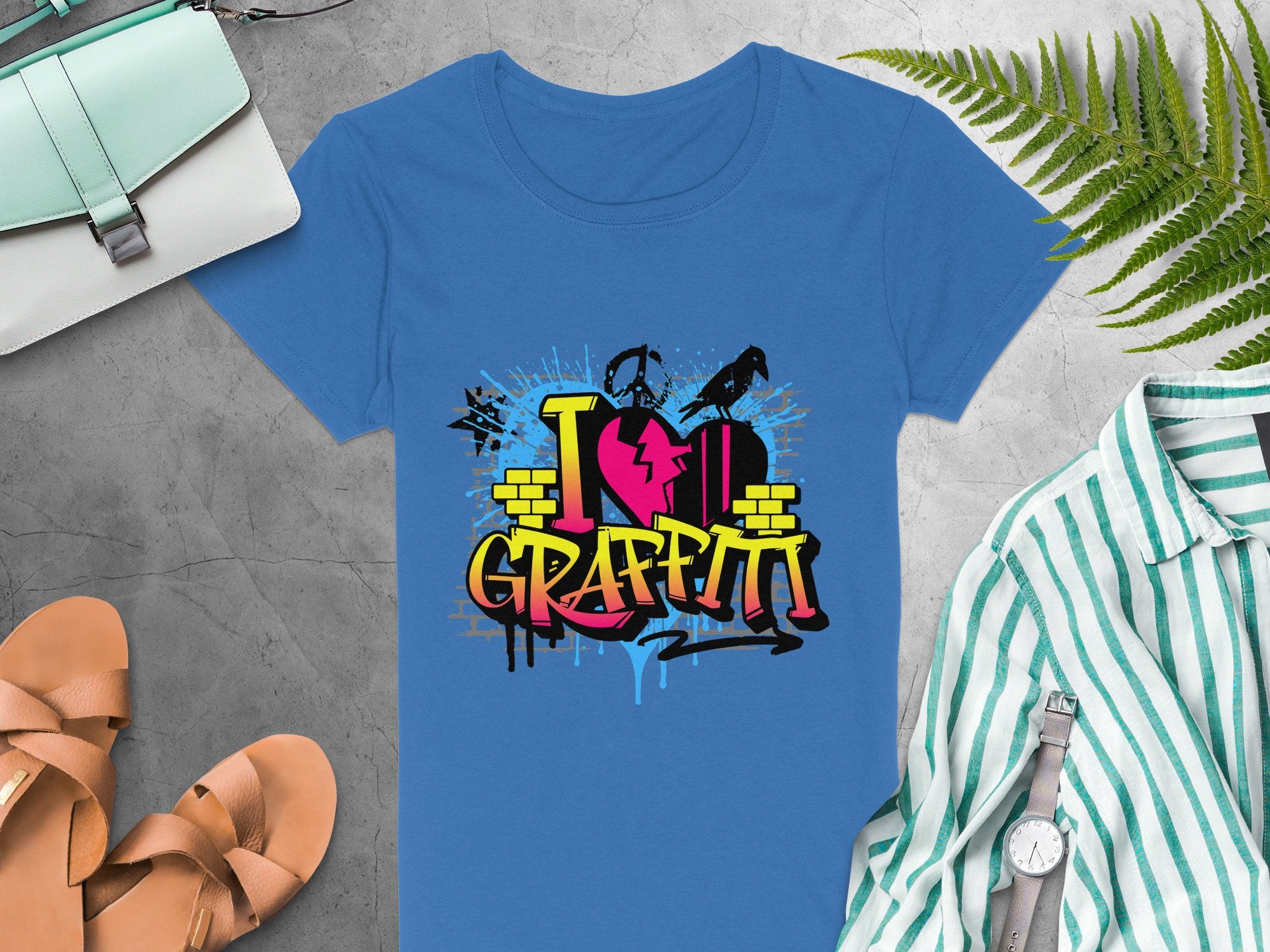 A Garment Graphics blue T-shirt with a vibrant I Love Graffiti street art design is displayed on a gray surface, accompanied by urban accents like sandals, a striped shirt, a wristwatch, a green purse, and green leaves.