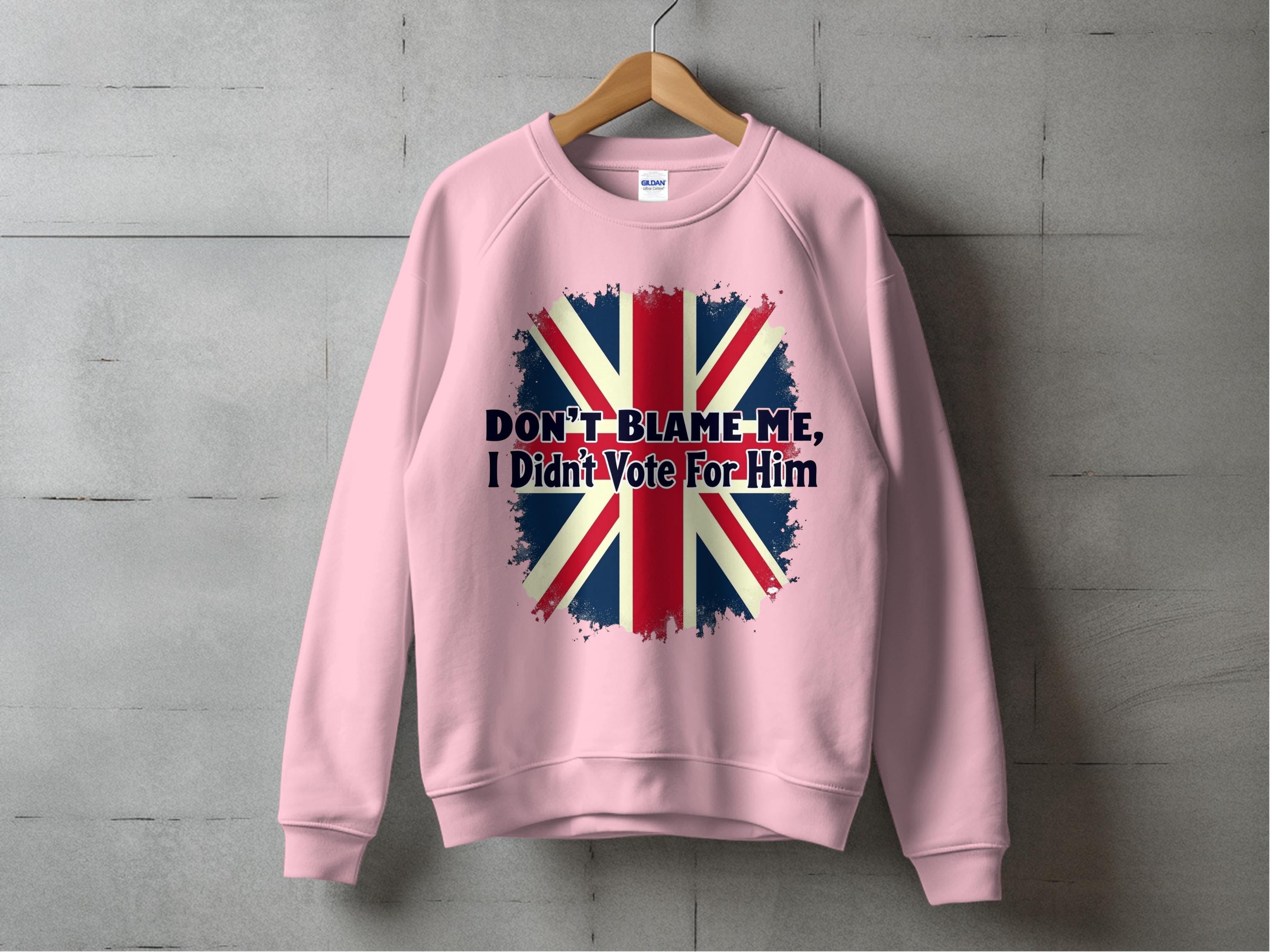 A cozy pink sweatshirt by Garment Graphics hangs on a wooden hanger against a concrete wall, featuring a bold, distressed Union Jack design with DONT BLAME ME, I Didnt Vote For Him printed over the flag.