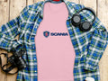 A soft pink Scania Logo T-shirt by Garment Graphics rests on a wooden surface, surrounded by a green and blue plaid shirt, black headphones, a camera, and a white mug with a lid—a charming ensemble of Scania merchandise.