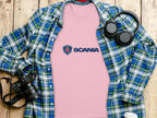 A soft pink Scania Logo T-shirt by Garment Graphics rests on a wooden surface, surrounded by a green and blue plaid shirt, black headphones, a camera, and a white mug with a lid—a charming ensemble of Scania merchandise.