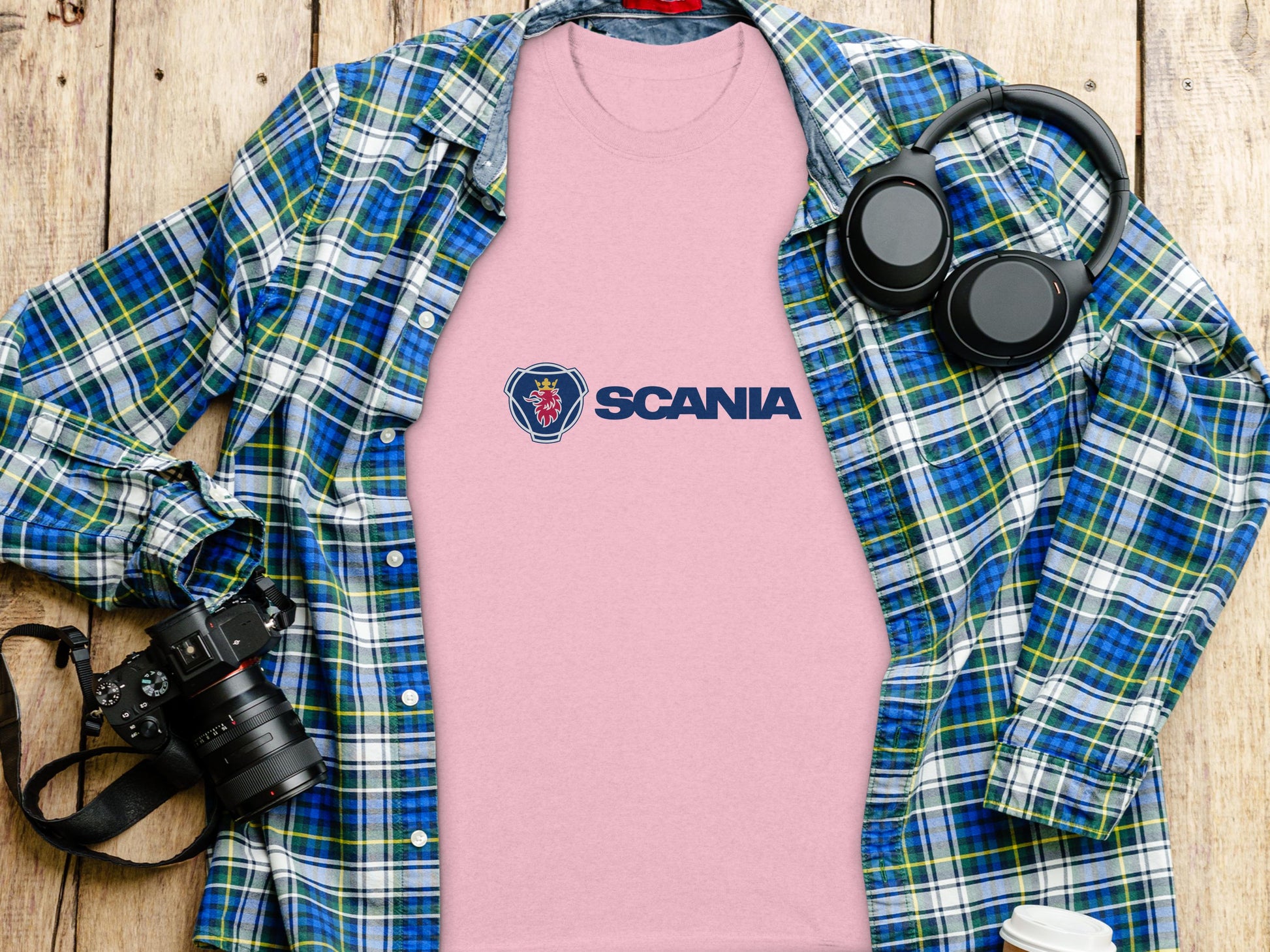 A soft pink Scania Logo T-shirt by Garment Graphics rests on a wooden surface, surrounded by a green and blue plaid shirt, black headphones, a camera, and a white mug with a lid—a charming ensemble of Scania merchandise.