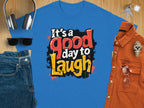A Garment Graphics Its a Good Day to Laugh blue t-shirt, featuring a vibrant black, red, and yellow message, is creatively displayed with gray headphones, sunglasses, jeans, keys, a brown watch, and an orange button-up shirt on a wooden surface.
